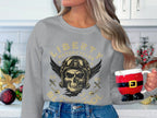 A person with long blonde hair embracing the biker look dons a Garment Graphics gray sweatshirt featuring a skull design, holding a Santa Claus mug. They stand before kitchen cabinets adorned with holiday decorations, merging coziness and edginess.