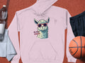 A pink unisex hoodie by Garment Graphics features a cartoon llama in sunglasses with the text No Prob-Llama. Its laid on a brown surface alongside essentials like a water bottle, black shorts, a basketball, and a light blue towel.