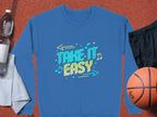 A blue unisex sweatshirt from Garment Graphics features Take It Easy with colorful graphics, like musical notes and arrows. Styled flat with a basketball, black shorts, a water bottle, and a towel nearby.