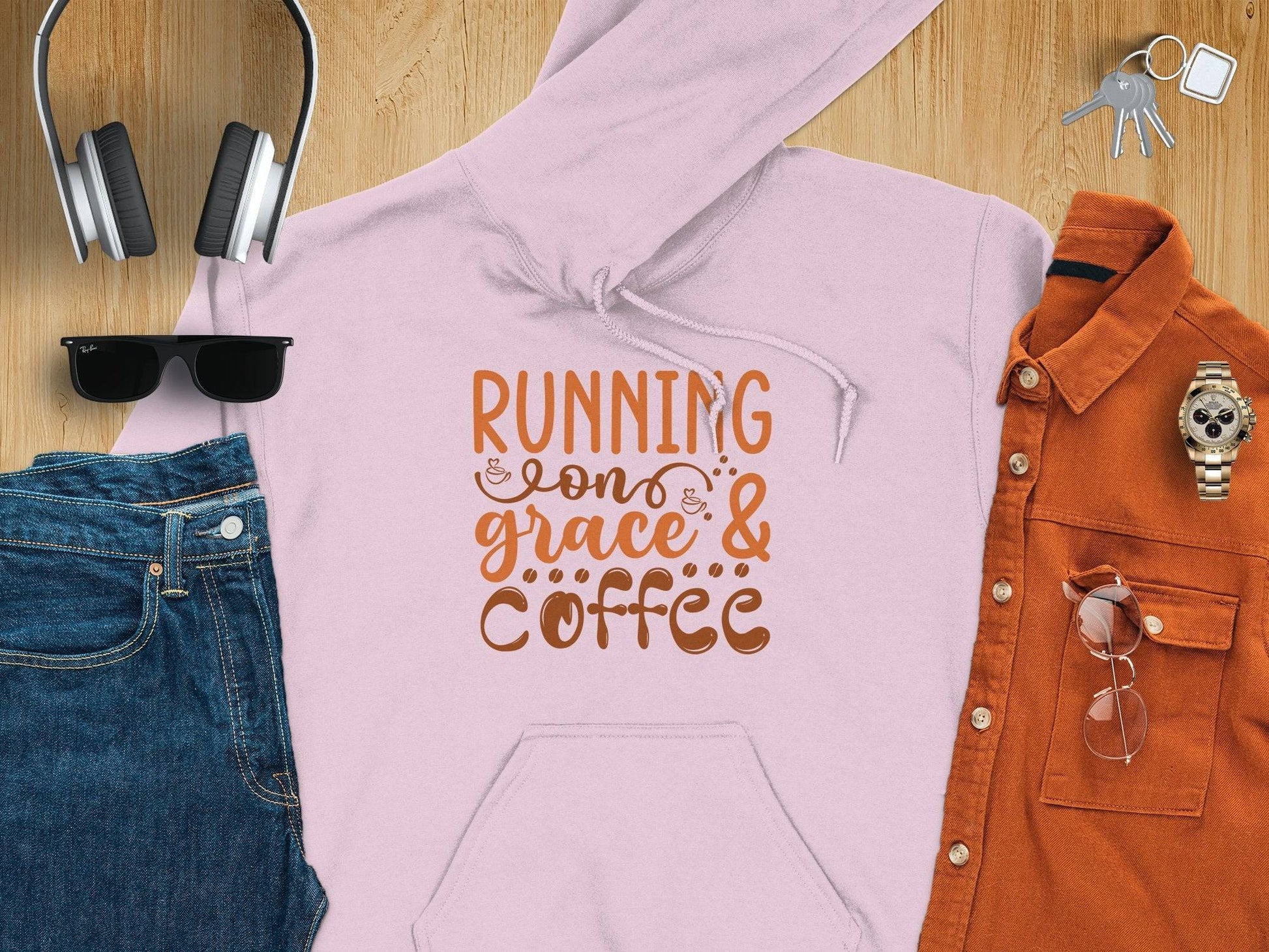 A flat lay features the Running on Grace and Coffee hoodie by Garment Graphics, made of medium-heavy fabric for a comfy classic fit. Surrounding the pink hoodie are black headphones, sunglasses, denim pants, a brown shirt, a wristwatch, clear glasses, and keys.