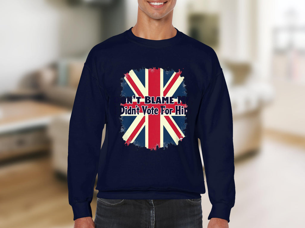 A person is wearing a unisex navy blue Garment Graphics sweatshirt with the UK flag and text: Dont Blame Me I Didnt Vote For Him, set against a softly blurred indoor background.