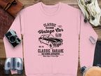 Garment Graphics pink sweatshirt showcases a vintage car graphic with Classic Vintage Car Club, Classic Garage text. Styled on wood, its paired with a camera, binoculars, and leather bag—ideal for vintage car enthusiasts.