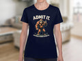 A person wears a navy Garment Graphics t-shirt with a cartoon Bigfoot and the phrase ADMIT IT. Life would be boring without me, set against a blurred indoor background, enhancing the playful vibe of this Admit It Bigfoot tee.