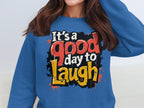 Someone is wearing a Garment Graphics sweatshirt in medium-heavy fabric, featuring the text Its a good day to Laugh in colorful, bold letters. The unisex sizing offers comfort for everyone, with vibrant orange, red, and yellow splatter designs in the background.