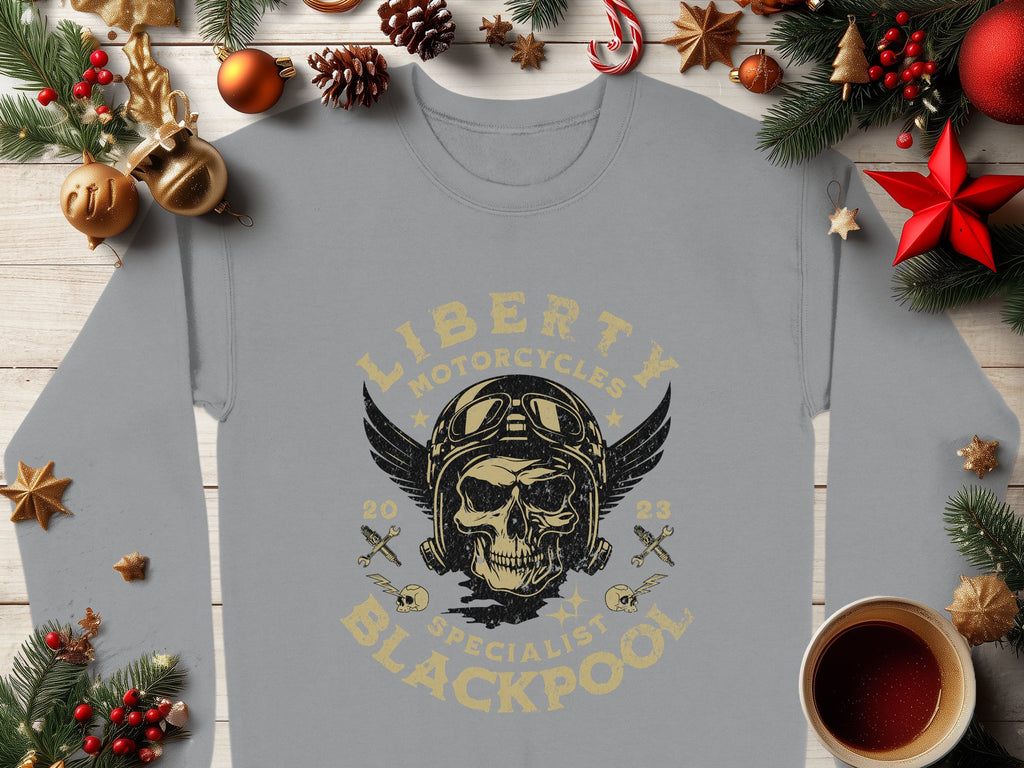 Garment Graphics offers a gray sweatshirt with Liberty Motorcycles Specialist Blackpool design, featuring a skull with goggles and wings. The backdrop includes pine branches, red stars, and a steaming cup on wood, merging unique style with Blackpool 2023 flair.