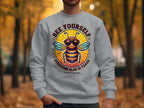 A person is wearing a Garment Graphics Summer Edition sweatshirt with a bee in sunglasses and the text Bee Yourself, Everyone Else is Taken. The unisex sizing sweatshirt is set against a wooded area with stunning autumn foliage.
