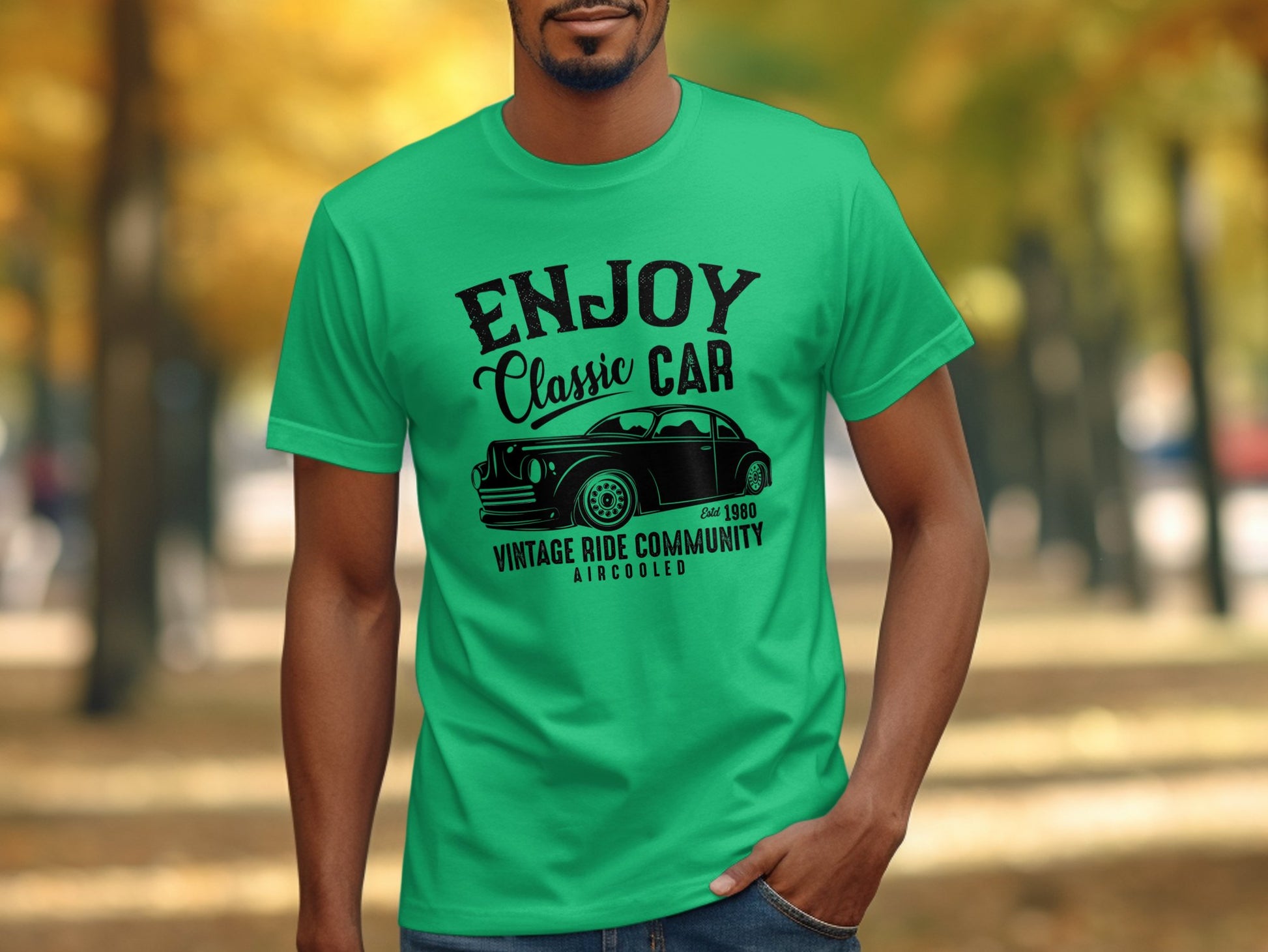 A person in a Garment Graphics green T-shirt featuring a classic car and the words Enjoy Classic Car, Vintage Ride Community, Estd 1980, Aircooled, stands against blurred trees and fallen leaves—a perfect scene for vintage ride enthusiasts.