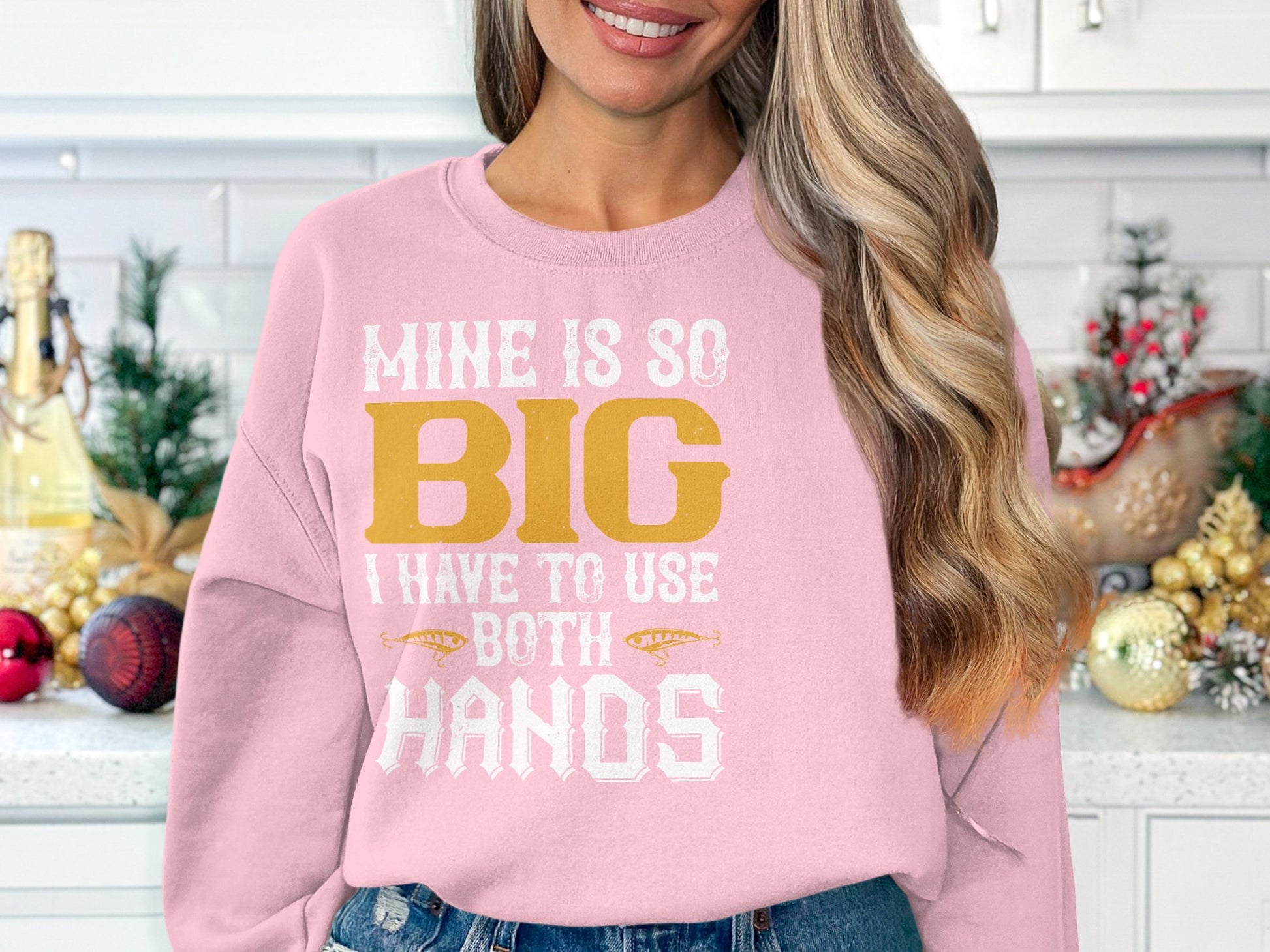 In a festive kitchen, a person wears a Garment Graphics pink sweatshirt with the bold phrase MINE IS SO BIG I HAVE TO USE BOTH HANDS. Their long, wavy hair enhances the unique look.