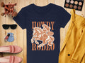 A navy Rodeo T-shirt by Garment Graphics showcases Western charm with a cowboy design and phrases Howdy Rodeo and Not My First Rodeo. Styled for women, its matched with a mustard jacket, makeup, sunglasses, pink shoes, and nail polish on a wooden surface.