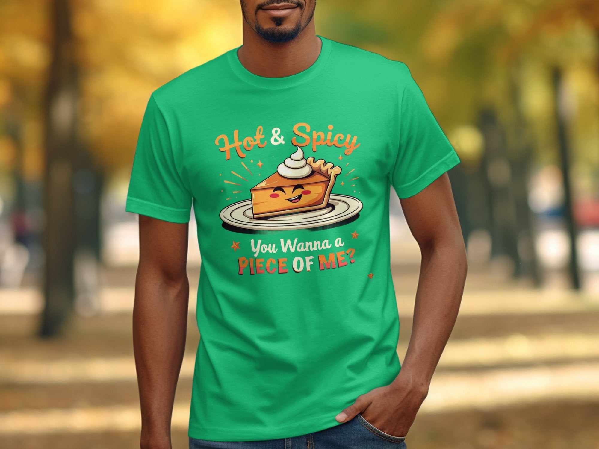 In a park with blurred autumn trees, someone wears a green Garment Graphics t-shirt featuring a winking pie slice topped with cream, boldly stating Hot & Spicy, You Wanna a Piece of Me? Perfect for grabbing attention and suitable for the whole family.