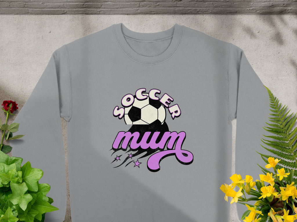 Garment Graphics presents a gray sweatshirt with Soccer Mum in purple and black, adorned with a soccer ball above the text and stars with swooshes below. Styled for vintage flair, it rests on stone while green and yellow plants enliven the scene.