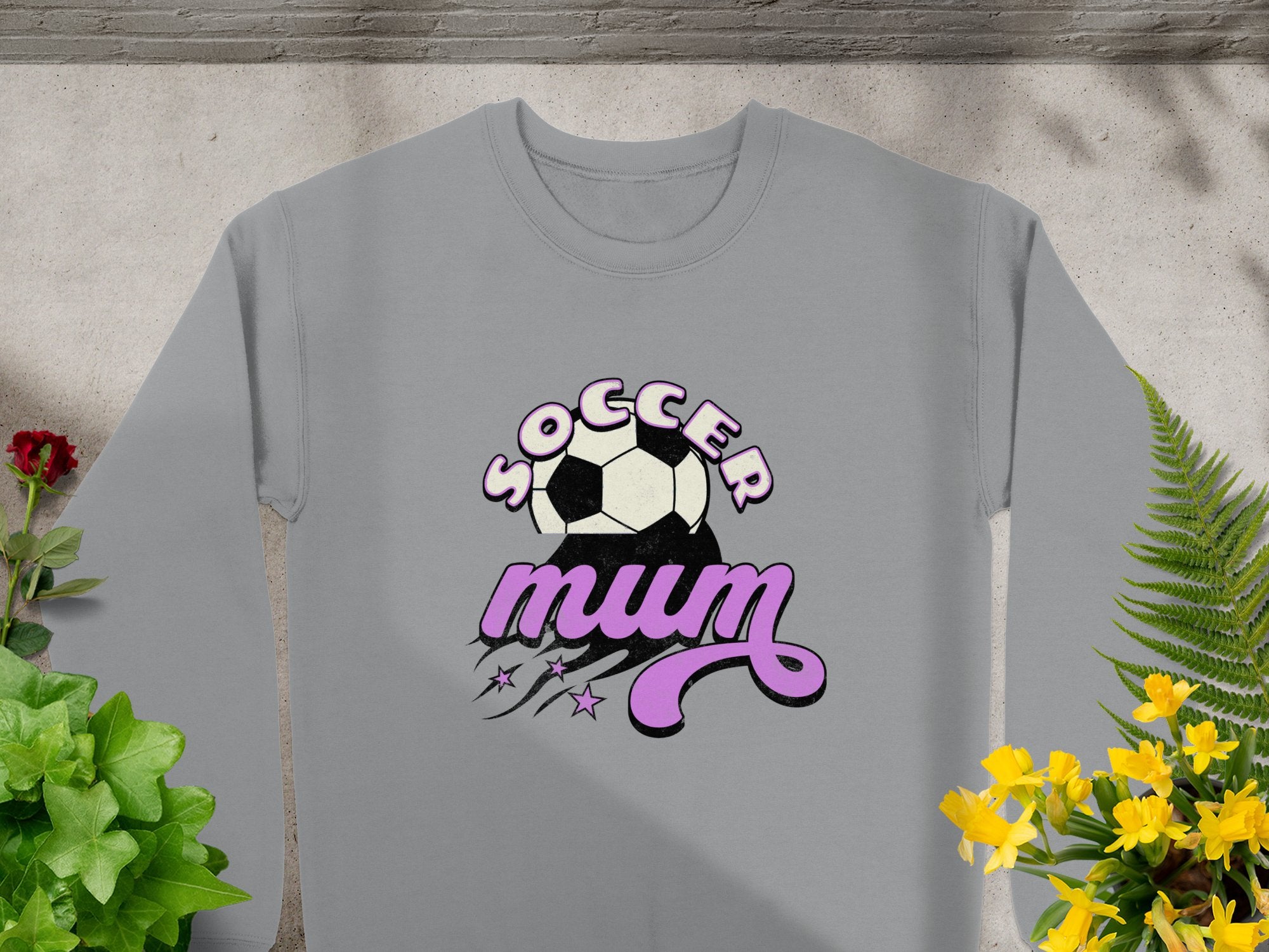 Garment Graphics presents a gray sweatshirt with Soccer Mum in purple and black, adorned with a soccer ball above the text and stars with swooshes below. Styled for vintage flair, it rests on stone while green and yellow plants enliven the scene.