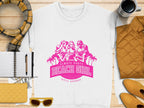A Garment Graphics white t-shirt with a pink design featuring three women, palm trees, and Always Party Beach Girl. Its stylishly displayed on wood, surrounded by a scarf, notebook, mug, jacket, wallet, and sunglasses.