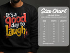 A person wears a black Garment Graphics sweatshirt featuring the vibrant red and yellow text Its a Good Day to Laugh. Next to them is a size chart for unisex Gildan 18000 sweatshirts, detailing sizes S to 5XL with precise width and length measurements.
