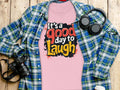 Garment Graphics presents a semi-fitted pink t-shirt with Its a good day to laugh text, resting on wood. A blue and green plaid shirt complements it. Nearby, a camera, headphones, and takeaway coffee cup create an upbeat vibe.