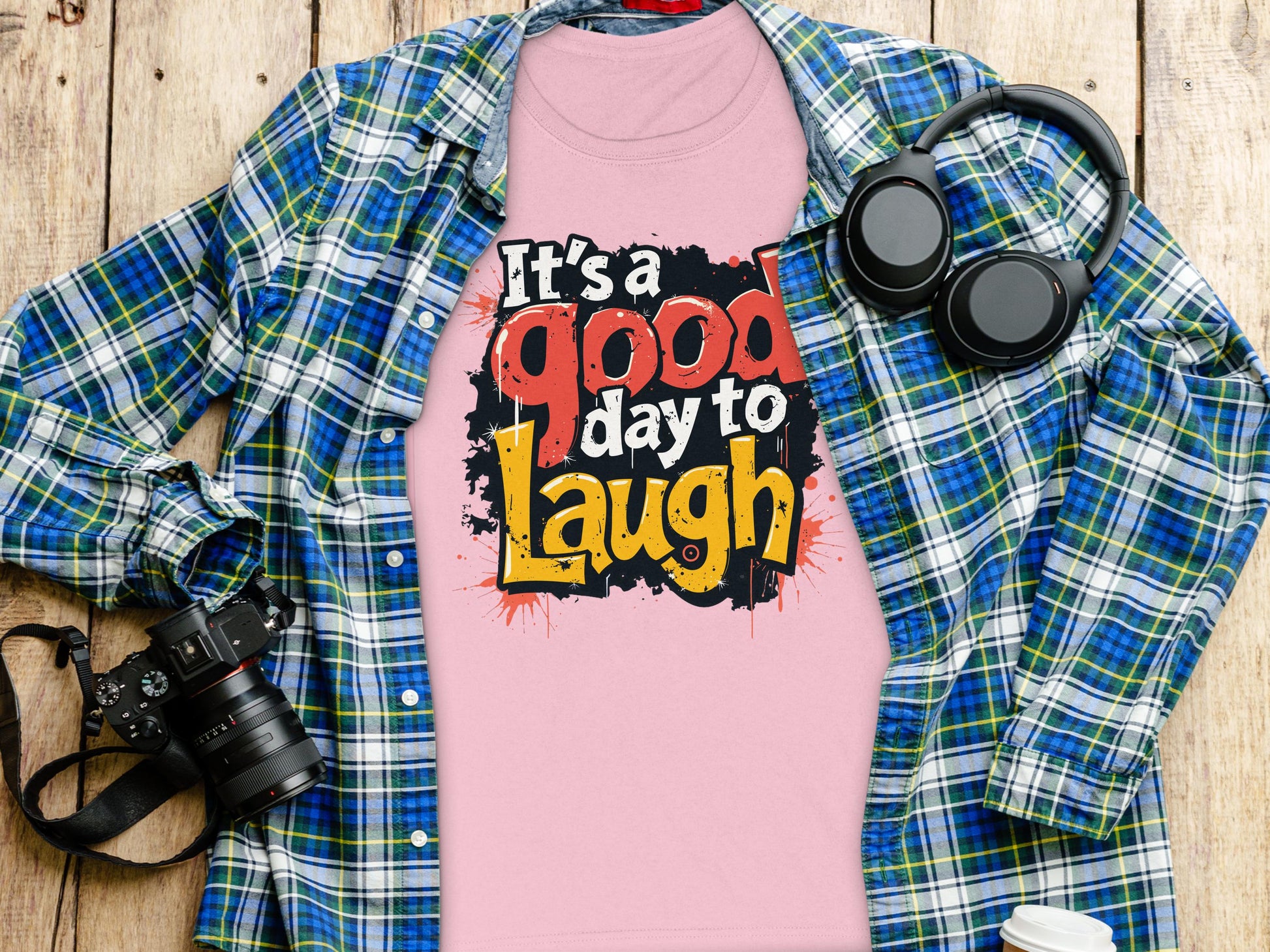 Garment Graphics presents a semi-fitted pink t-shirt with Its a good day to laugh text, resting on wood. A blue and green plaid shirt complements it. Nearby, a camera, headphones, and takeaway coffee cup create an upbeat vibe.
