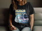 A person wears a black Garment Graphics T-shirt featuring a retro surf design: a classic car with surfboards, waves, palm trees, and the bold yellow text BEACH VIBES. Seated and shown from shoulders down, it impeccably captures timeless beach vibes.