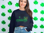 A person with long hair smiles in a Garment Graphics festive sweatshirt featuring a green truck and shamrocks, reading Happy St. Patricks Day. Green shamrock decorations are visible in the background, enhancing the celebratory spirit.