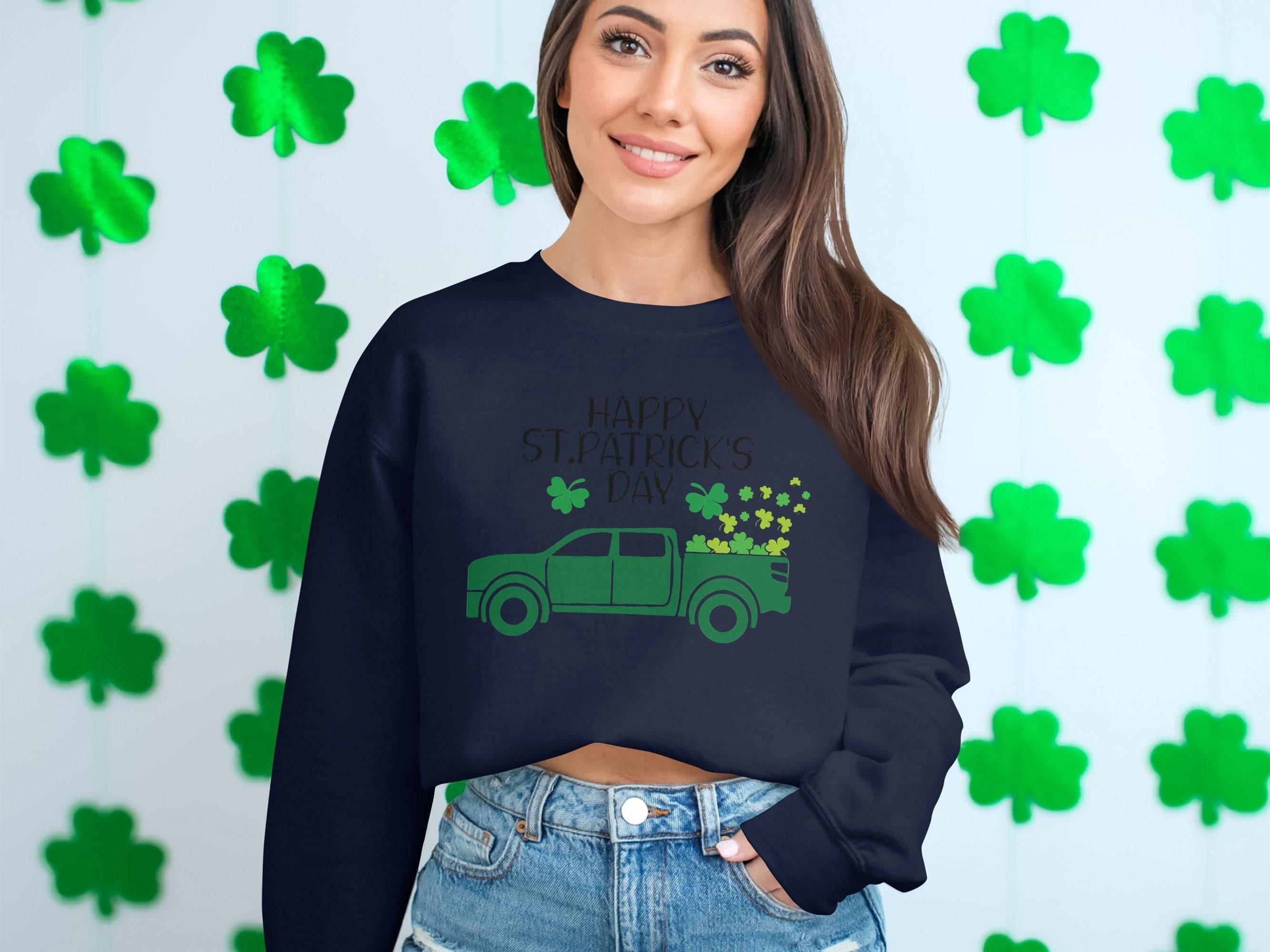 A person with long hair smiles in a Garment Graphics festive sweatshirt featuring a green truck and shamrocks, reading Happy St. Patricks Day. Green shamrock decorations are visible in the background, enhancing the celebratory spirit.