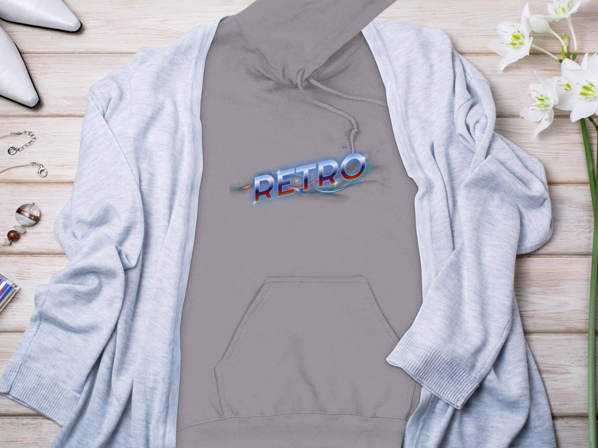 A gray hoodie featuring a vibrant 80s-style Retro graphic by Garment Graphics lays flat on wood, paired with a light gray cardigan. White pointed shoes, jewelry, perfume, and white flowers embellish this chic ensemble.