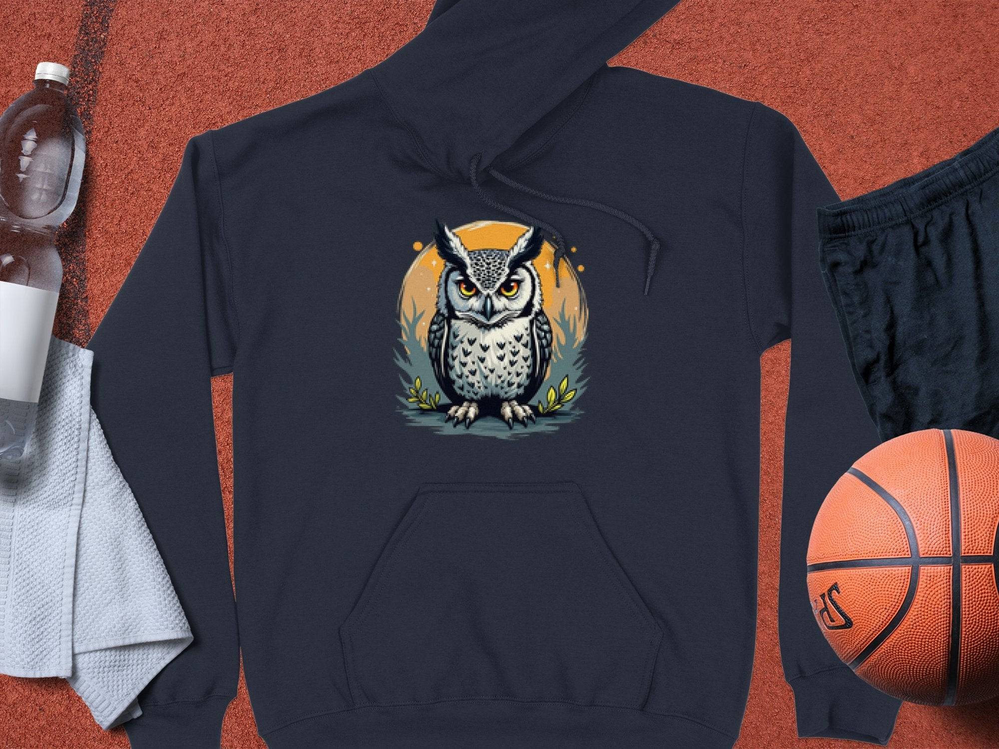 A medium-heavy fabric hoodie by Garment Graphics features an owl design on a dark gray backdrop with an orange circle, laid on a textured surface alongside a plastic water bottle, white towel, black shorts, and an orange basketball.