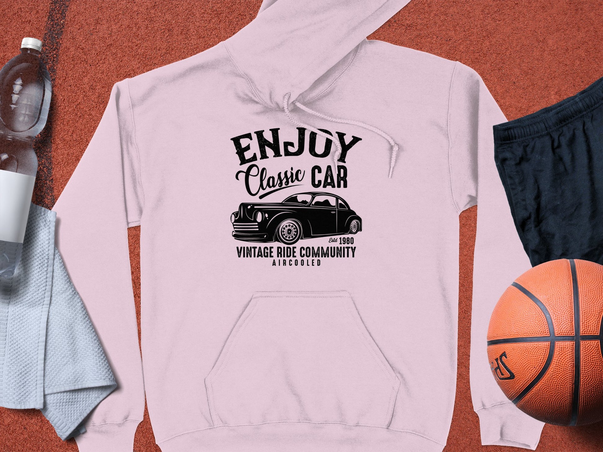 Garment Graphics presents a light pink vintage car hoodie, featuring Enjoy Classic Car and a retro car graphic, symbolizing the community spirit with tags like Vintage Ride Community and Aircooled. Its paired with a water bottle, black shorts, a white towel, and an orange basketball on brown surface.