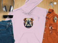 Flat lay of a Garment Graphics light pink hoodie with a bold bulldog print, paired with jeans, an orange button-up shirt, headphones, sunglasses, keys, and a watch on a wooden surface. A spiked collar adds edge to this statement piece.