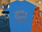 A blue classic fit T-shirt from Garment Graphics, featuring THIS DAY IS Powered BY Coffee, lies flat on a wooden surface alongside jeans, sunglasses, headphones, a brown shirt, and a watch, exuding positivity.