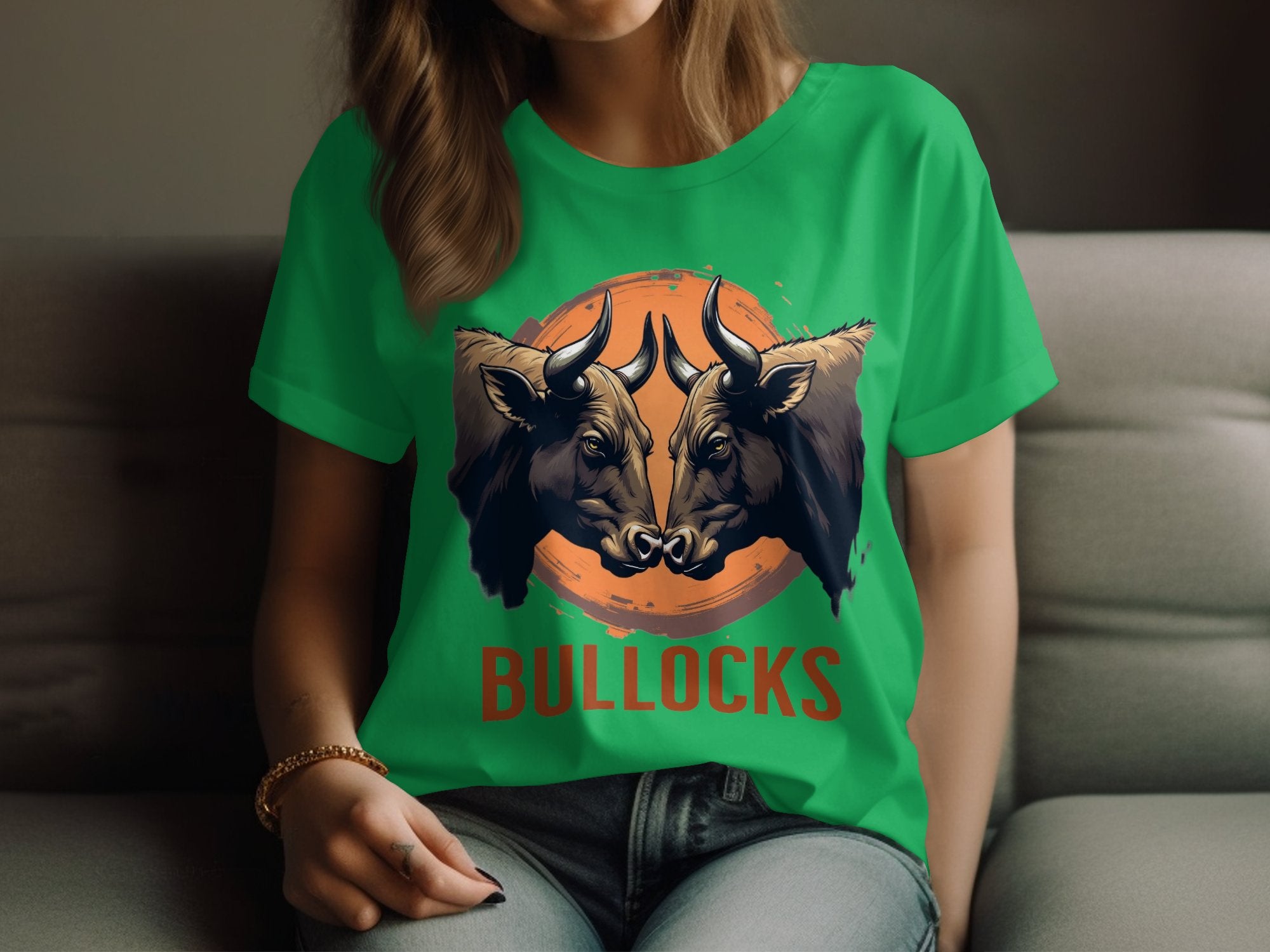 A person wearing a green t-shirt from Garment Graphics, showcasing bold graphics of two bulls facing each other with BULLOCKS printed beneath, sits on a gray couch. Their right hand casually rests on their lap, enhancing the laid-back style.