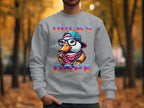 A person dons a Garment Graphics gray sweatshirt showcasing a cartoon duck with glasses, cap, and scarf alongside DUCKS DOPE in bold letters. Against a backdrop of forest and fallen leaves, this design embodies Brave Cool Swag Sweatshirts style, offering medium-heavy fabric comfort.