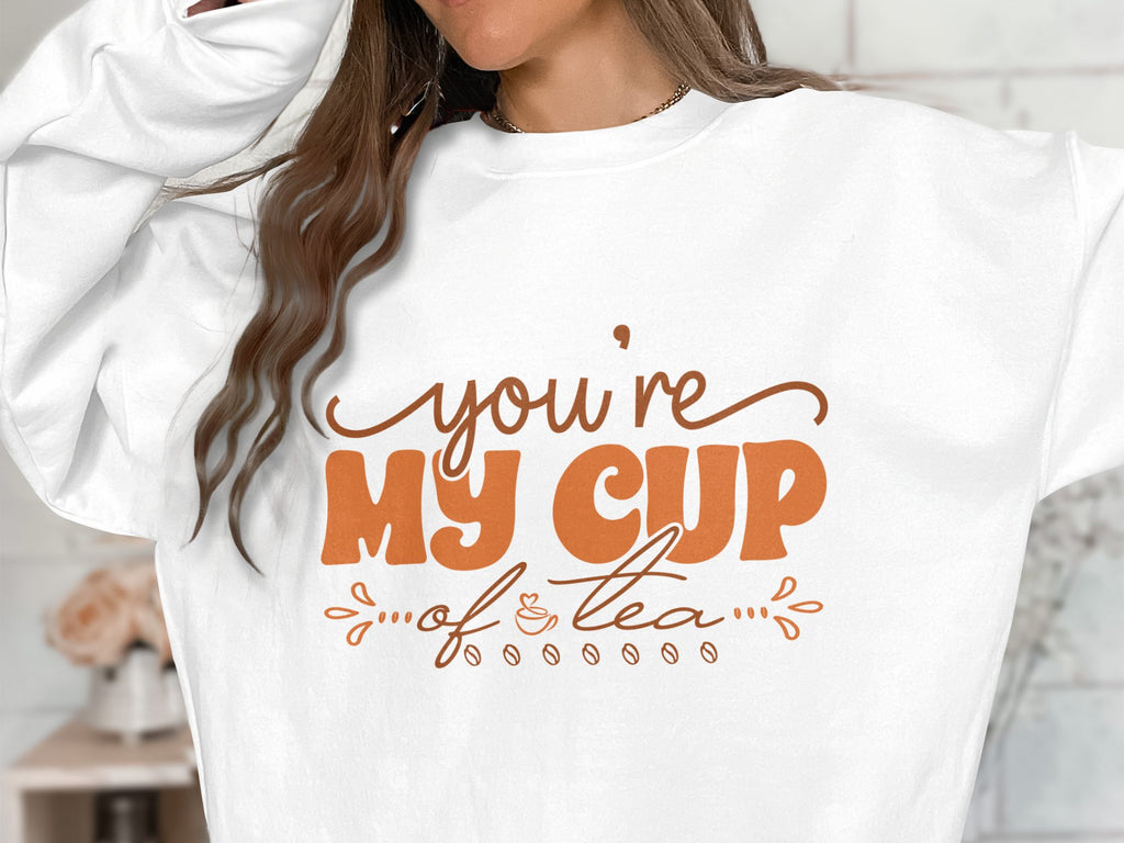 A person wears a cozy Garment Graphics sweatshirt with Youre My Cup of Tea elegantly printed in brown letters. Their long hair partially obscures their face, while the unisex sizing of the sweatshirt adds to its universal charm.