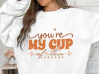 A person wears a cozy Garment Graphics sweatshirt with Youre My Cup of Tea elegantly printed in brown letters. Their long hair partially obscures their face, while the unisex sizing of the sweatshirt adds to its universal charm.