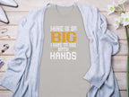 A humorous Garment Graphics gray t-shirt with MINE IS SO BIG I HAVE TO USE BOTH HANDS in orange and white text, perfect for fishing enthusiasts, is displayed on a wooden surface surrounded by a light blue cardigan, white flowers, rings, and a necklace.