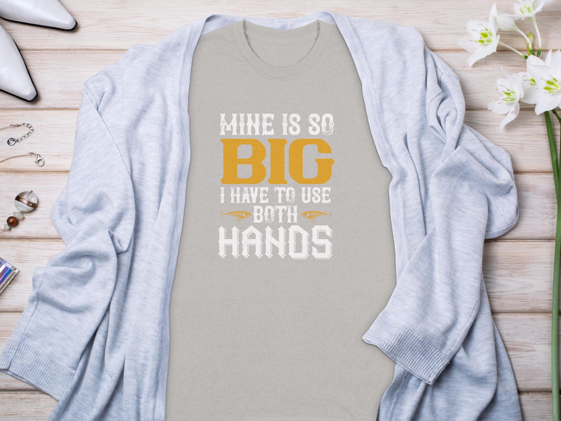 A humorous Garment Graphics gray t-shirt with MINE IS SO BIG I HAVE TO USE BOTH HANDS in orange and white text, perfect for fishing enthusiasts, is displayed on a wooden surface surrounded by a light blue cardigan, white flowers, rings, and a necklace.