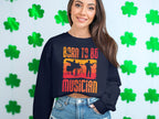 A smiling woman wears a black BORN TO BE MUSICIAN sweatshirt by Garment Graphics featuring silhouettes of two musicians. This unisex design is ideal for music lovers. She poses against a white wall with green shamrock cutouts.