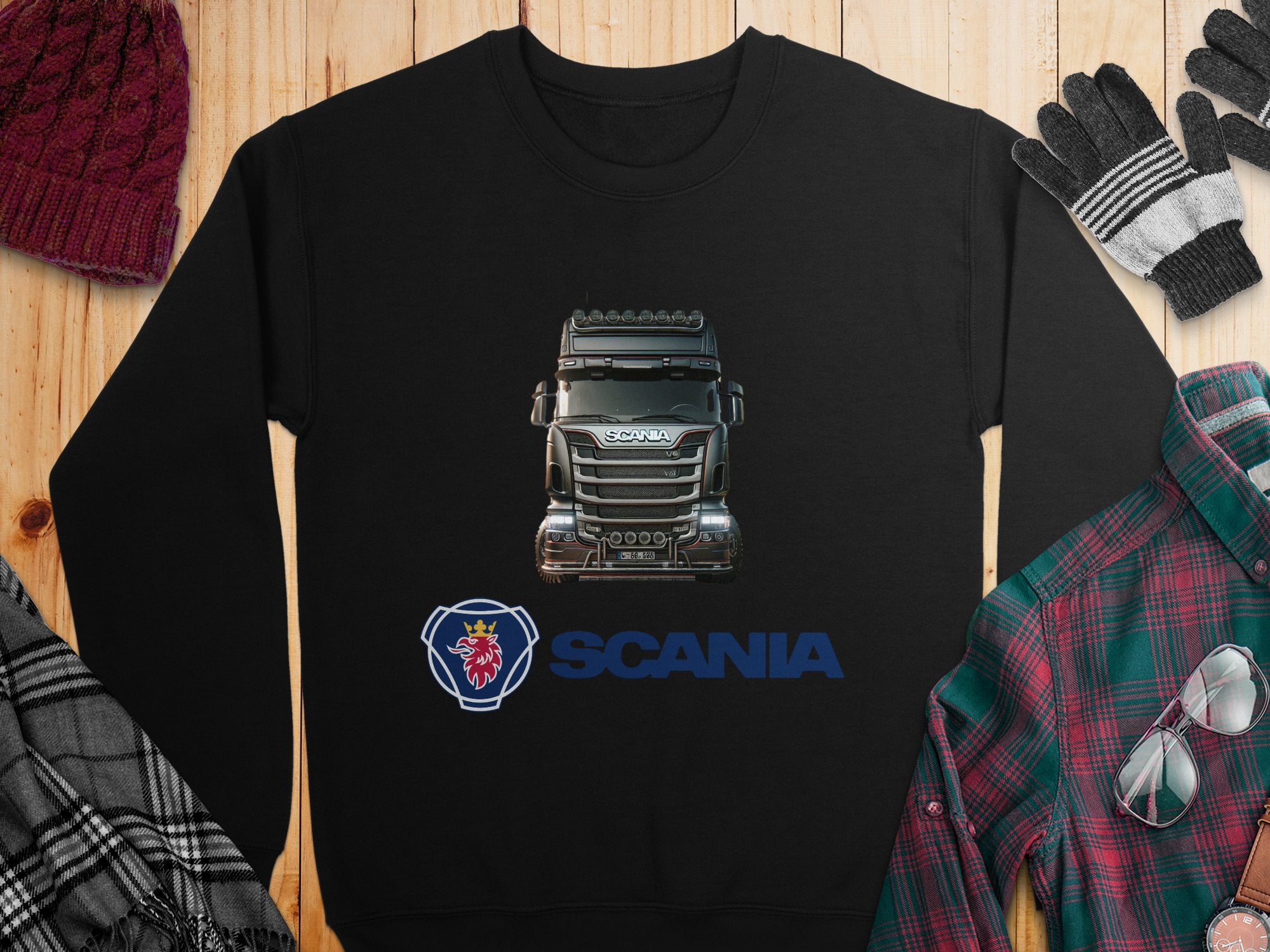 A black graphic crewneck sweatshirt by Garment Graphics, featuring a Scania truck and logo, lies on a wooden surface. Its ideal for truck fans. Nearby are a plaid shirt, hat, gloves, and scarf.