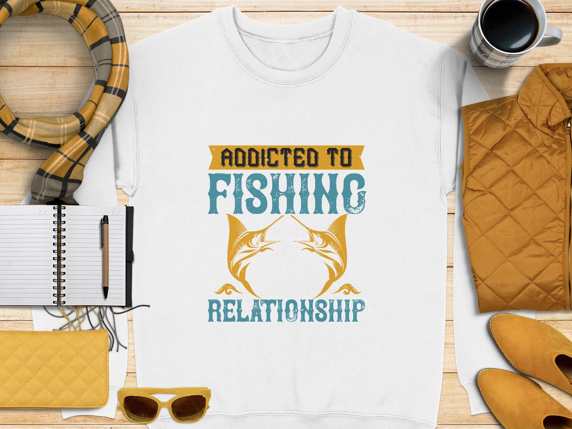 Garment Graphics offers a white unisex T-shirt featuring Addicted to Fishing Relationship with two stylized fish. Complemented by a yellow scarf, quilted jacket, notebook and pen, coffee cup, yellow clutch, and sunglasses—ideal for any fishing enthusiasts casual day out.