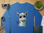 A blue Garment Graphics No Prob-Llama sweatshirt, featuring a llama in red sunglasses, is displayed on a wooden surface among a camera, lens, small bag, kiwi bowl, book, and map. It offers unisex sizing.