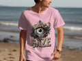 A person walks along the beach in a classic fit pink T-shirt from Garment Graphics, featuring a large ornate eye illustration with jewels and pearls. GAZE is boldly printed beneath the eye, showcasing unique designs as the ocean shimmers in the background.