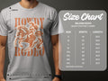 Someone is wearing a gray Garment Graphics T-Shirt, showcasing a cowboy riding a horse and Howdy Rodeo in orange. Ideal for enthusiasts, it comes with a Gildan 5000 size chart, detailing sizes S-5XL with width and length measurements in inches.