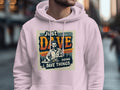 A person sports a light pink Garment Graphics hoodie made from medium-heavy fabric, featuring a vintage-style man and the text Just Dave Doing Dave Things against a city street backdrop with blurred pedestrians.