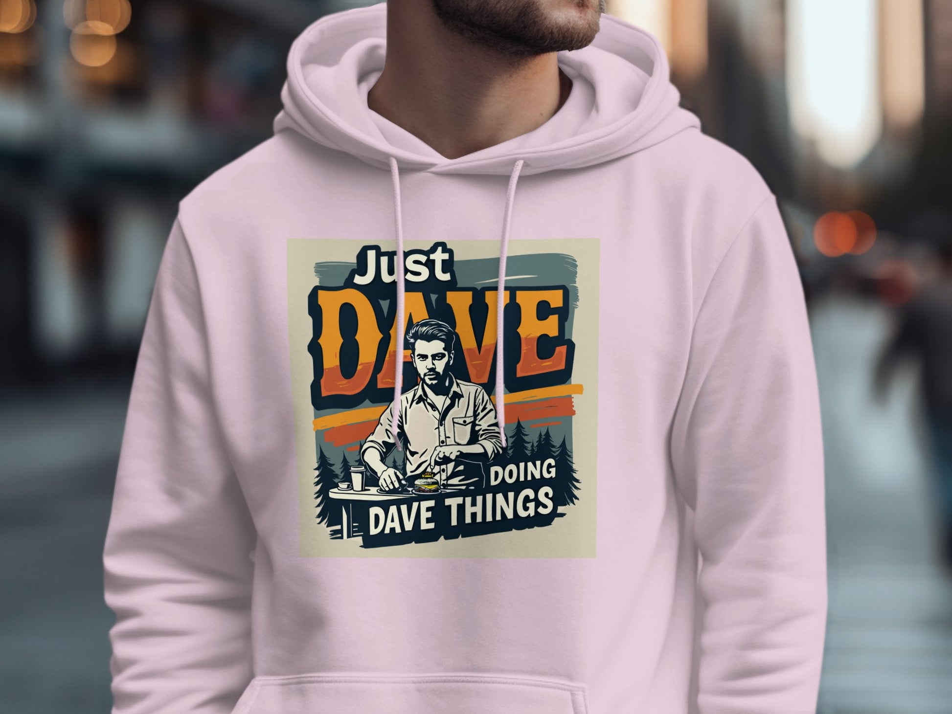 A person sports a light pink Garment Graphics hoodie made from medium-heavy fabric, featuring a vintage-style man and the text Just Dave Doing Dave Things against a city street backdrop with blurred pedestrians.