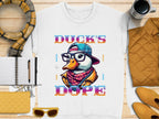 Make a bold fashion statement with Garment Graphics t-shirt featuring a cartoon duck wearing glasses, headphones, and a hat. The words DUCKS DOPE are surrounded by wood accents, a scarf, notebook, pen, coffee cup, quilted jacket, and yellow wallet and sunglasses.