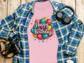 A vibrant, 100% cotton pink T-shirt by Garment Graphics with Happy Birthday and colorful balloons sits on wood next to a blue-green plaid shirt. A camera, headphones, and a white disposable cup with a black lid add to the lively celebration atmosphere.