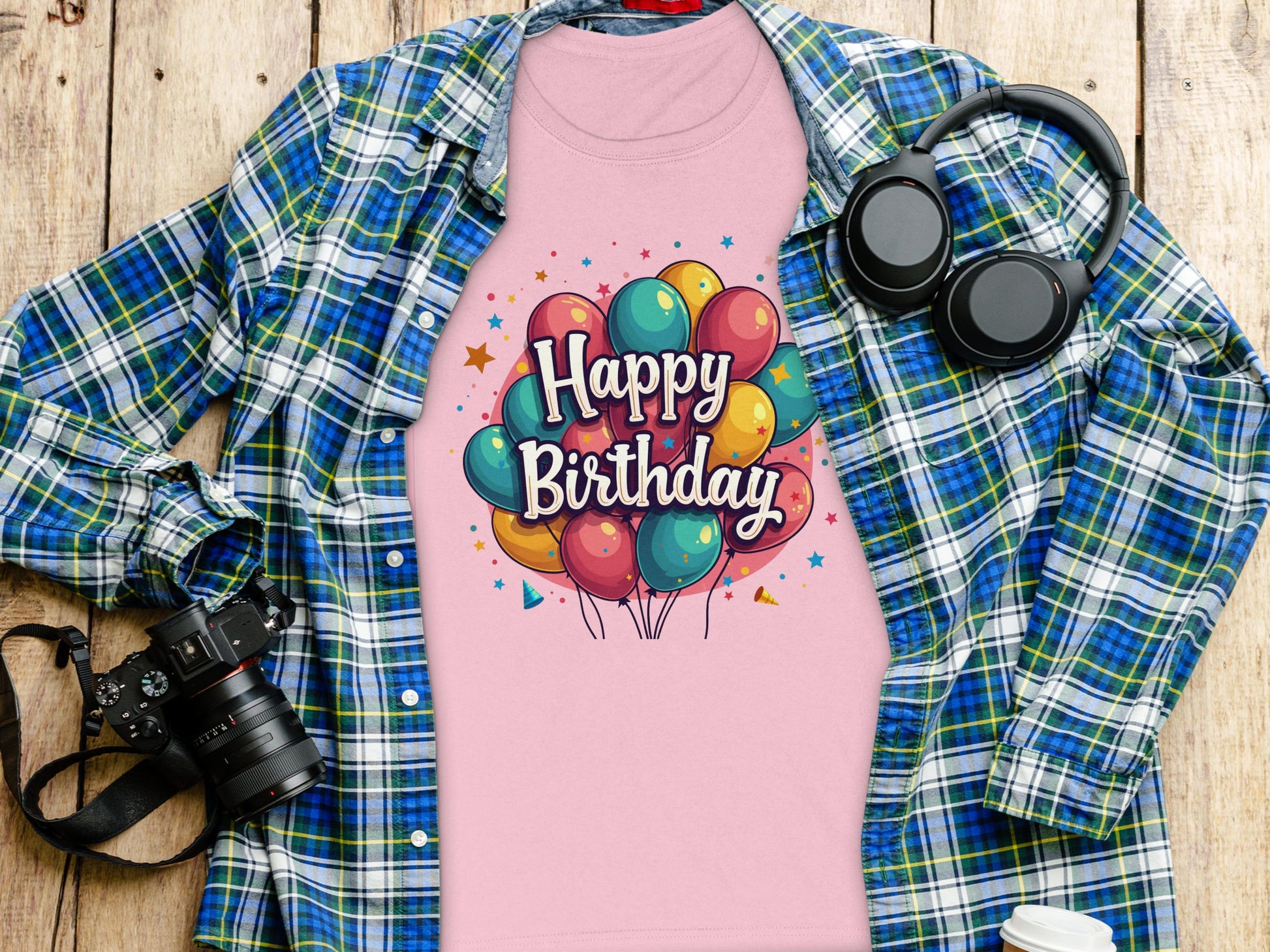 A vibrant, 100% cotton pink T-shirt by Garment Graphics with Happy Birthday and colorful balloons sits on wood next to a blue-green plaid shirt. A camera, headphones, and a white disposable cup with a black lid add to the lively celebration atmosphere.