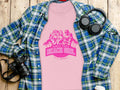 A pink Always Party Beach Girl t-shirt with fun illustrations is on a wooden surface, surrounded by a blue plaid shirt, headphones, and a camera, capturing the carefree vibe of Beach Girl T-Shirts by Garment Graphics.
