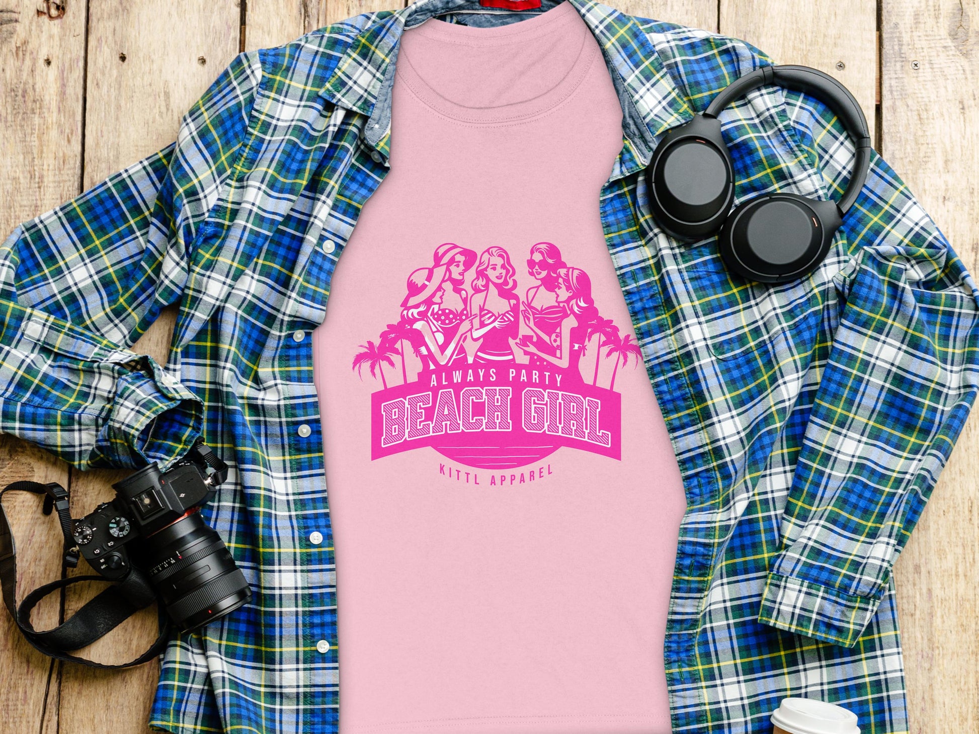 A pink Always Party Beach Girl t-shirt with fun illustrations is on a wooden surface, surrounded by a blue plaid shirt, headphones, and a camera, capturing the carefree vibe of Beach Girl T-Shirts by Garment Graphics.