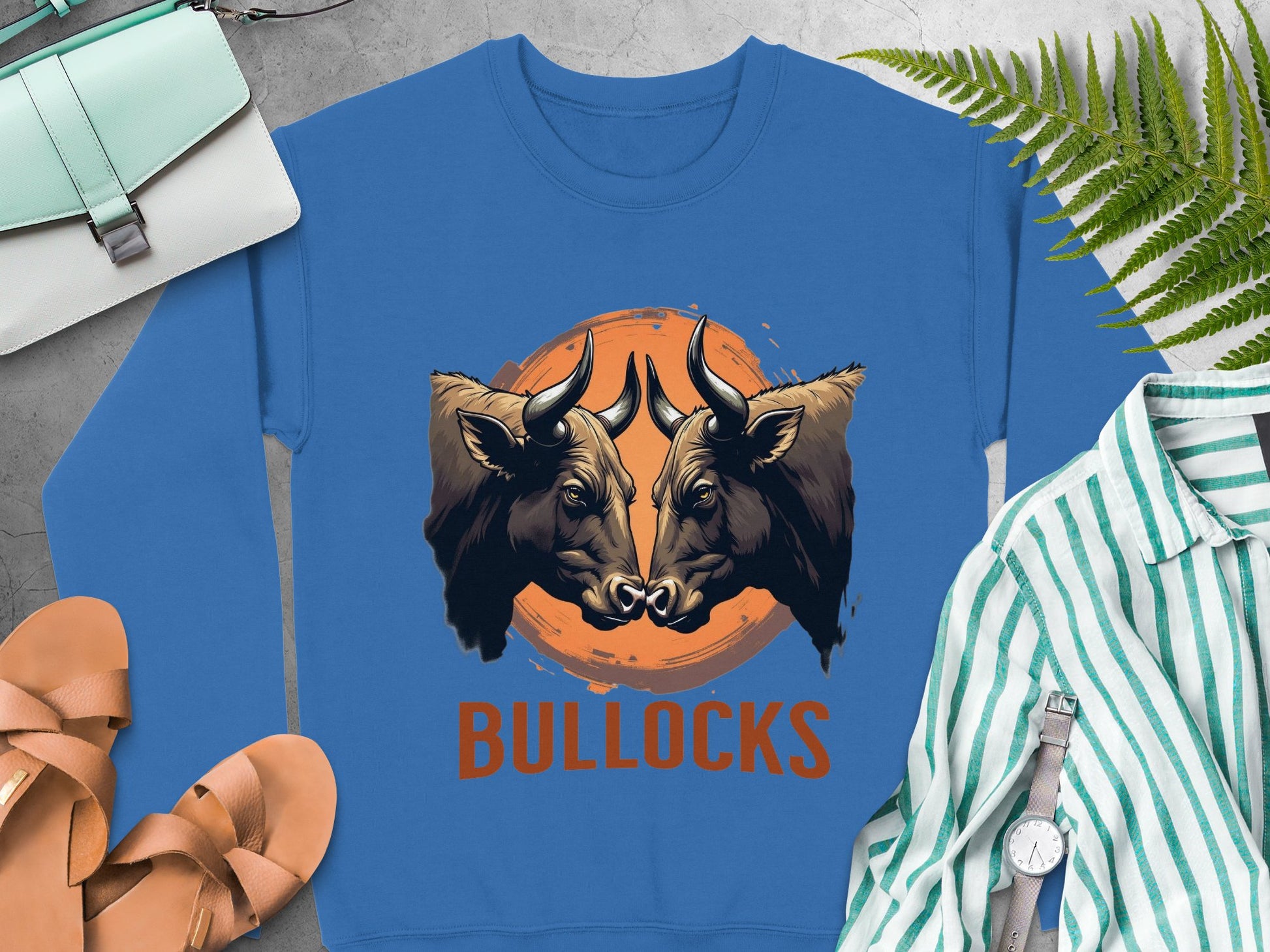 A blue, loose-fit sweatshirt from Garment Graphics features a striking graphic with two bulls locking horns over an orange circle, captioned BULLOCKS. Its styled on a surface with a bag, sandals, striped shirt, and vibrant ferns.
