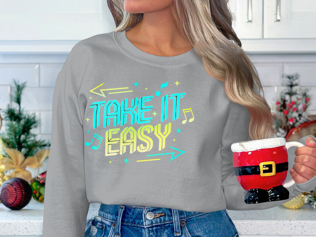 A person with long hair holds a Santa-themed mug, feeling cozy in a Garment Graphics take-it-easy graphic sweatshirt. This unisex piece is perfect for casual wear. Festive Christmas decorations on the kitchen counter enhance the holiday ambiance.