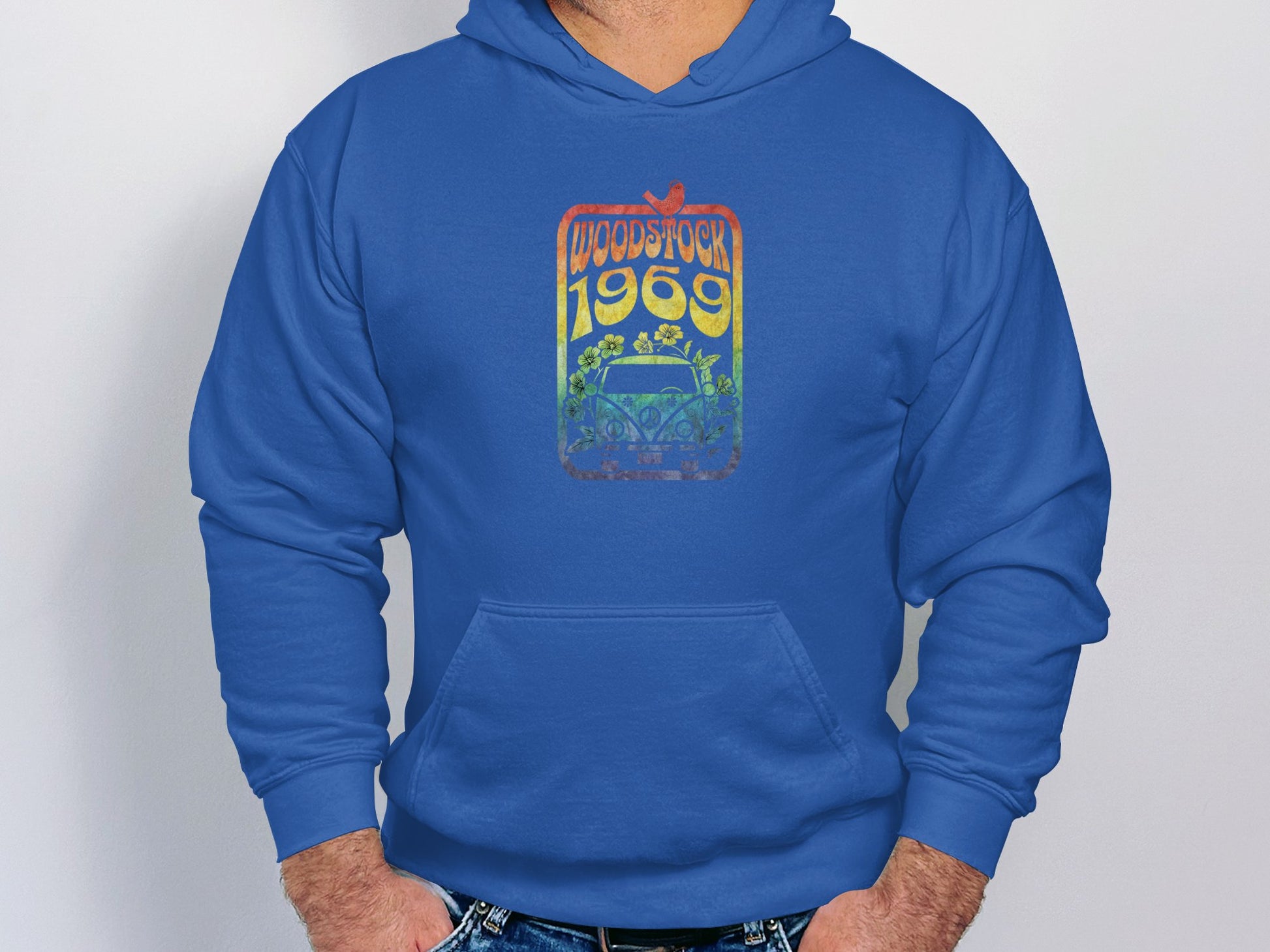 A person wearing a Garment Graphics retro blue hoodie featuring a vibrant Woodstock 1969 design with flowers and a car stands against a light gray background, hands in pockets.